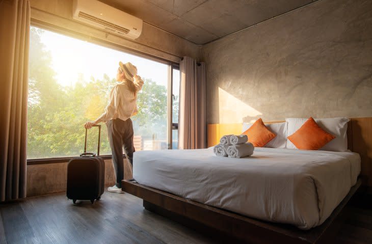 A woman standing next to her suitcase and looking out the window of her sunny hotel room.